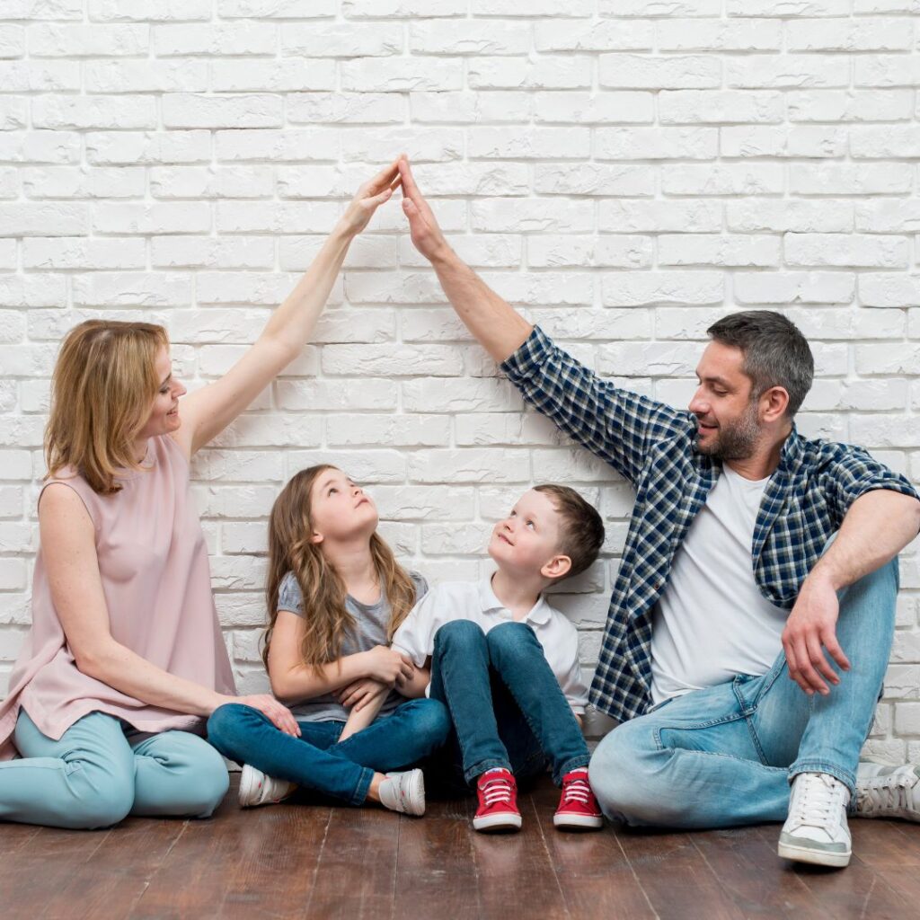 A family sitting against a white brick wall with the mother and father holding their hands to make a "roof" above their son and daughter.